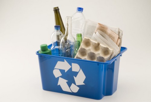 A blue plastic recycling container with a white recycling symbol on the front, filled with various types of waste including clear and green glass bottles, an empty plastic water bottle, a partially folded newspaper, and a cardboard egg carton, situated against a plain light background. The bottles vary in size and shape, with some capped and others open, and the newspaper appears crumpled. The container's rectangular shape and sturdy design are typical for waste collection, and the mixture of materials indicates it is used for mixed recycling or general rubbish collection, relevant to rubbish removal services in Seven Kings, Essex. The environment seems to be outside or a well-lit indoor setting, with the focus solely on the container and its contents, emphasizing sorting and disposal of waste materials as part of local waste management activities provided by Commercial Waste Removal Seven Kings.