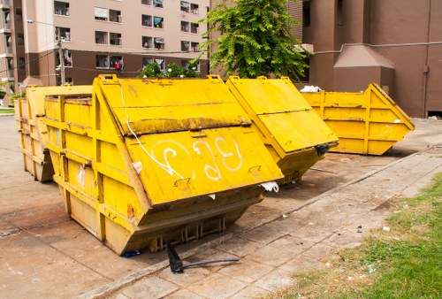 A large yellow metal skip bin with reinforced side panels and a slightly weathered finish, positioned on a paved concrete surface in an urban residential area. The bin is angled with its open top facing towards the right side of the image, and features white graffiti markings on its front panel. Behind the skip, there are two additional yellow skip bins aligned along the pavement, with their sides visible. In the background, multi-storey residential blocks with brown brick facades, balconies, and windows are partially obscured by a lush green tree. The surrounding environment includes a small patch of grass and a concrete driveway, indicating a typical city street setting. The overall scene is illuminated by natural daylight, emphasizing the weathered texture of the metal and the vibrant colour of the skips, which are indicative of waste collection activities often undertaken by services like Commercial Waste Removal Seven Kings in the area.