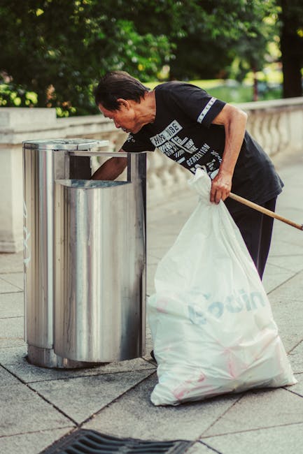 An outdoor scene depicting a large collection of mixed rubbish and waste materials on a dirt ground, including numerous black and white plastic bags, cardboard boxes, oversized wooden spools, discarde